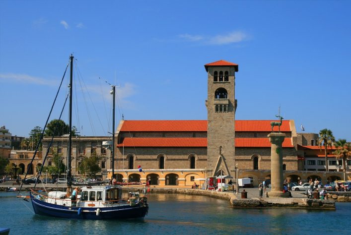 Hafenpanorama mit Segelboot vor historischem Gebäude mit Turm, erinnert an Standort des Koloss, eines Weltwunders der Antike.