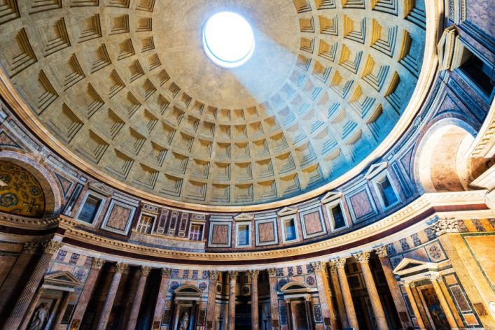 Im Pantheon von Rom fällt Licht durch eine kreisrunde Öffnung in der Kuppel. Die verzierte Wand zeigt Marmorsäulen und kunstvolle Reliefs. Das architektonische Meisterwerk ist beeindruckend groß.