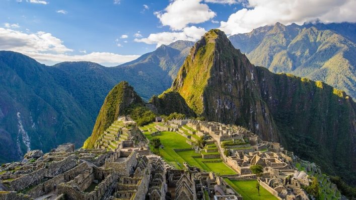 Inmitten grüner Berge liegt die antike Inka-Stadt Machu Picchu, deren steinerne Ruinen sich weitläufig ausbreiten. Im Hintergrund erheben sich majestätische Gipfel unter einem klaren Himmel.