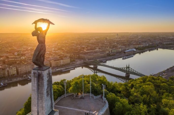 Die Freiheitsstatue auf dem Gellértberg bei Sonnenuntergang, im Hintergrund der Blick auf die Donau, die Szabadság-Brücke und die Stadt Budapest. Tipps: Fantastischer Aussichtspunkt für Fotos und Spaziergänge.