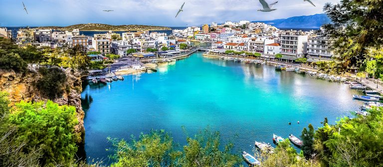 Türkisfarbener See in Agios Nikolaos auf Kreta, umgeben von beigen und weißen Gebäuden, Booten am Ufer und Möwen im blauen Himmel.
