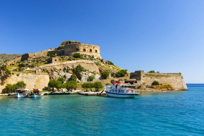 Festungsruine auf Spinalonga vor Kretas Küste, umgeben von klarem, blauem Wasser, unter strahlend blauem Himmel. Vorne Schiffe am Anlegeplatz.