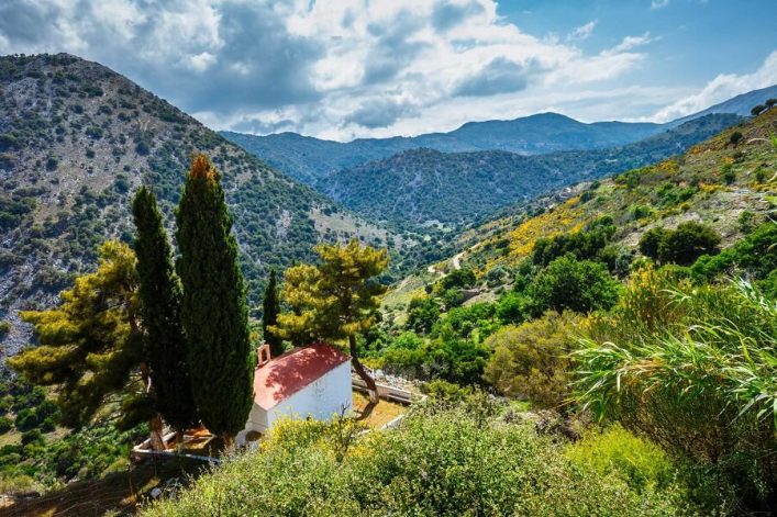 Eine kleine Kirche mit rotem Dach, umgeben von Zypressen, inmitten des grünen Lassithi-Plateaus auf Kreta, mit Bergen im Hintergrund.