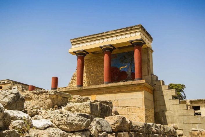 Antike rote Säulen und Fresken des Palasts von Knossos unter klarem blauem Himmel, umgeben von Ruinen, ein Wahrzeichen Kretas.
