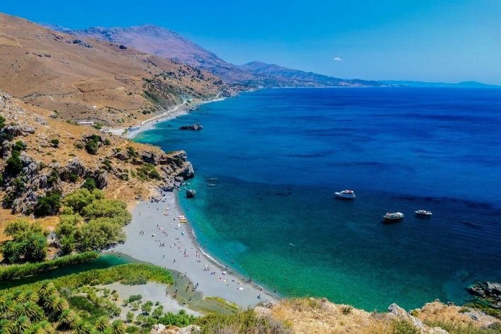 Das Bild zeigt den Preveli Beach. Eine Mischung aus Hügeln, grünen Pflanzen und dem hellgrauen Strand ist zu sehen. Ganz rechts auf dem Bild sieht man das türkisblaue Meer und ein Schiff darauf.
