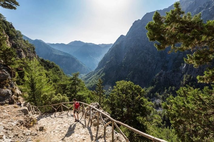 Ein Wanderweg führt an der Samaria-Schlucht auf Kreta entlang, umgeben von hohen Felswänden und grünen Bäumen. Ein Wanderer genießt die Aussicht.