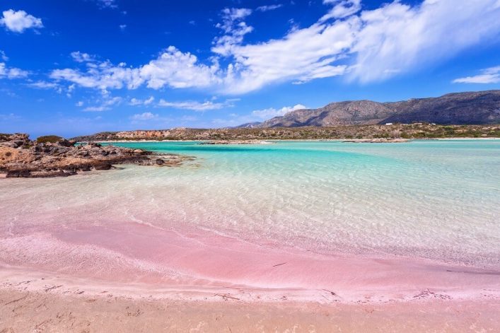 Rosa Sand trifft auf türkisblaues Meer unter einem blauen Himmel, während im Hintergrund karge Berge Griechenlands die Kulisse eines perfekten Urlaubs am Meer bilden.