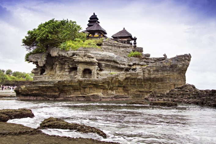 Der Tempel Tanah Lot thront auf einem Felsen, umgeben von Meerwasser. Links ist üppiges Grün, rechts steigt die Küste empor. Der Himmel ist bewölkt, das Meer brandet sanft an den Felsen.