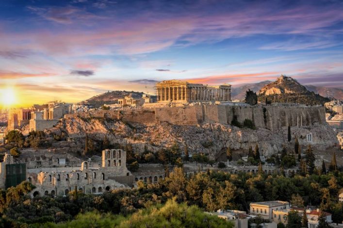 Sonnenuntergang über der Akropolis in Athen. Der Parthenon erhebt sich majestätisch auf dem Hügel, umgeben von Ruinen und grüner Vegetation. Im Hintergrund erstrahlt der Himmel in bunten Farben.