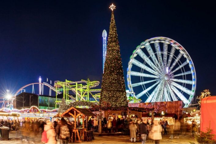 Beleuchteter Weihnachtsbaum in der Mitte, umgeben von belebten Marktständen. Rechts hinten ein großes, leuchtendes Riesenrad. Links leuchtet eine Achterbahn gegen den dunklen Nachthimmel.