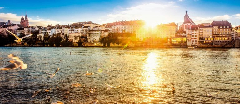 Blick auf Basel bei Sonnenuntergang, das Licht spiegelt sich im Rhein. Links erhebt sich der Basler Münster, rechts eine Kirche. Möwen fliegen über das Wasser, eine ruhige, friedliche Szene.
