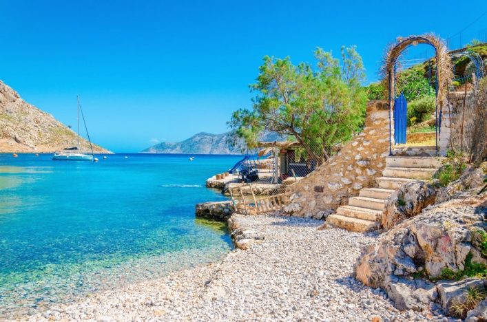 Kiesstrand mit türkisfarbenem Wasser, ein Segelboot links im Meer. Rechts Treppe aus Stein mit blauem Tor, von Pflanzen umgeben. Im Hintergrund Berge und blauer Himmel. Entspanntes Küstenambiente.