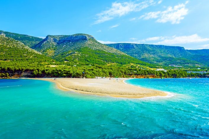 Strand mit goldenen Sand in Kroatien, türkisblaues Wasser auf beiden Seiten, umgeben von üppigen grünen Bergen im Hintergrund. Menschen genießen den sonnigen Tag auf dem Sand und im Wasser.