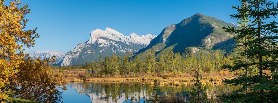 Majestätische Berge, von Schnee bedeckt, spiegeln sich in einem ruhigen See. Umgeben von herbstlichen Bäumen, erstreckt sich ein Nadelwald am Ufer. Der Himmel ist strahlend blau.