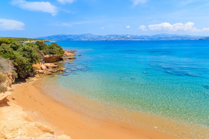 Sandstrand mit klarem, türkisfarbenem Wasser rechts, grünen Sträuchern links. Im Hintergrund schimmert das Meer unter einem strahlend blauen Himmel mit vereinzelten Wolken.