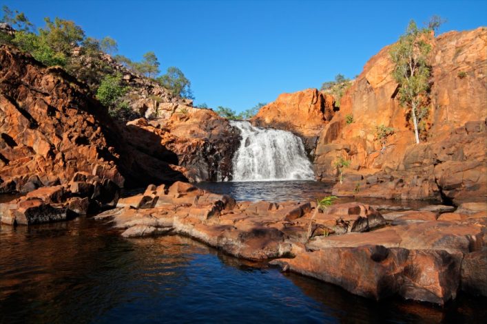 Wasserfall stürzt über rote Felsen in einen kleinen Fluss im Vordergrund. Dichte Vegetation mit Bäumen und Gebüsch auf den Felsen, klarer blauer Himmel im Hintergrund.