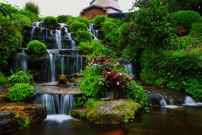Wasserfall plätschert über mehrere Felsstufen, umgeben von üppigem Grün und roten sowie rosa Blüten. Im Hintergrund ist ein Gebäude sichtbar, Bäume rahmen die Szene ein.