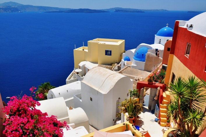 Weiße und bunte Häuser mit blauen Kuppeln am Hang einer Klippe in Santorini, Griechenland. Im Vordergrund pinke Bougainvillea, rechts Palmen. Im Hintergrund das tiefblaue Meer und ferne Inseln.