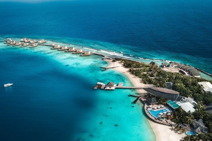 Luftaufnahme eines luxuriösen Inseldorfes auf den Malediven. Überwasserbungalows erstrecken sich in einer Reihe über türkisfarbenes Wasser, umgeben von Korallenriffen. Rechts Palmen und Sandstrände.