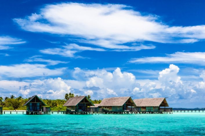Über türkisblauem Wasser reihen sich vier Holzbungalows auf Stelzen. Im Hintergrund erstreckt sich ein üppiger grüner Dschungel unter einem strahlend blauen Himmel mit weißen Wolken.