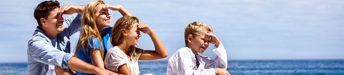 Vier Personen stehen nebeneinander und schauen mit erhobener Hand in die Ferne über das Meer. Der Himmel ist klar. Von links nach rechts: ein Mann, zwei Frauen und ein Junge. Alle lachen.