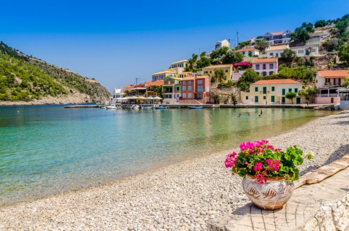 Klares türkisblaues Meer mit bunten Häusern am Hang im Hintergrund, Kiesstrand im Vordergrund mit einem Tontopf voller blühender roter und pinker Blumen. Menschen schwimmen im Wasser.