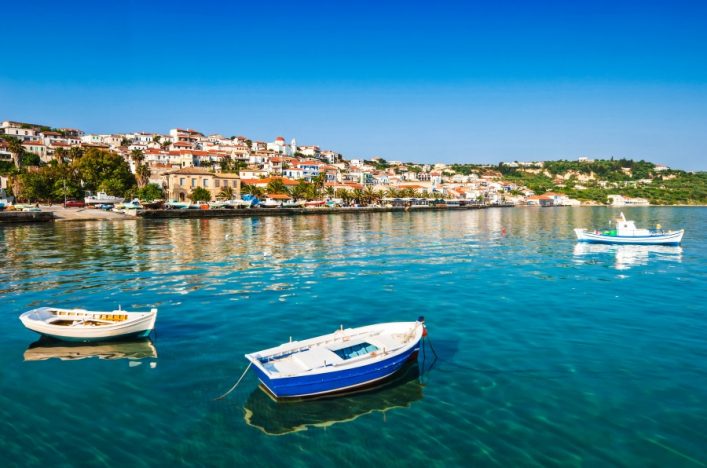 Klares, sonniges Wasser mit drei Booten im Vordergrund. Im Hintergrund bunte Häuser einer Küstenstadt auf einem grünen Hügel unter blauem Himmel.