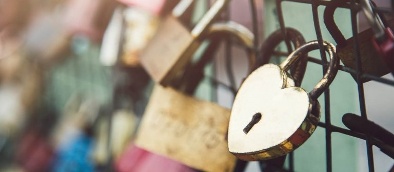 Verschiedenfarbige Liebesschlösser hängen an einem Zaun; ein herzförmiges Schloss in der Mitte im Fokus.