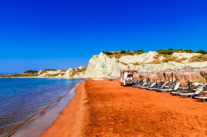 Roter Sandstrand mit strohbedeckten Sonnenliegen rechts. Türkisblaues Meer links, in der Ferne helle Felsklippen unter klarem blauem Himmel.