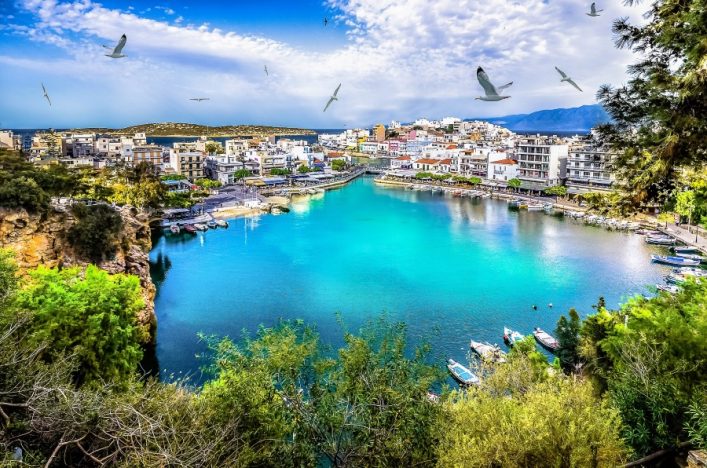 Das Bild zeigt in der Mitte ein türkisblaues Meer. Drumherum sind weiße Bauten und grüne Pflanzen zu sehen. Ganz oben ist ein blauer Himmel mit weißen Wolken und fliegenden Vögeln zu sehen.