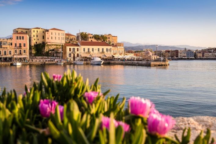 Blick auf den malerischen Hafen von Chania, Kreta, mit pastellfarbenen Häusern und Booten vor bergiger Kulisse, eingerahmt von rosa Blüten im Vordergrund.