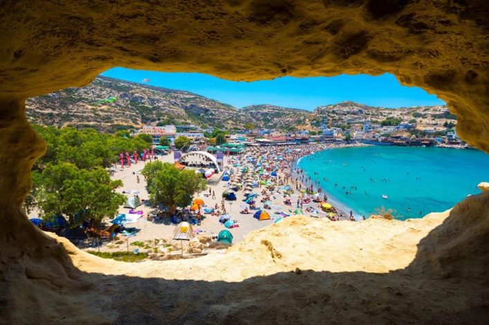 Blick durch eine Höhlenöffnung auf den belebten Matala Strand mit vielen Badegästen, bunten Schirmen und dem türkisfarbenen Meer auf Kreta.