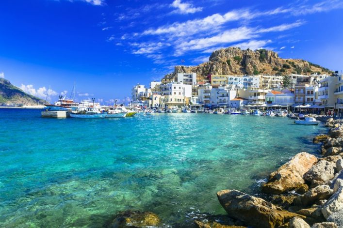 Klares türkisblaues Wasser im Vordergrund. Am Hafen liegen weiße Boote. Gebäude schmiegen sich an einen felsigen Hügel im Hintergrund unter einem strahlend blauen Himmel.