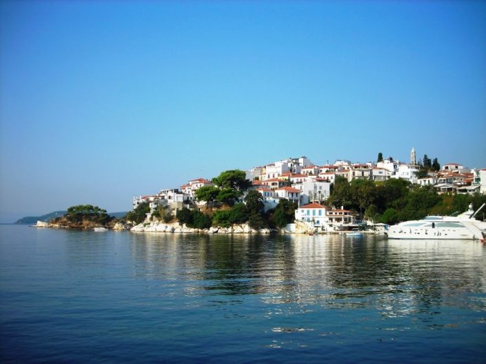 Insel mit weiß getünchten Häusern und roten Dächern, umgeben von üppigem Grün auf einer felsigen Küste. Im Vordergrund liegt ein großes weißes Boot im ruhigen, spiegelnden Wasser.
