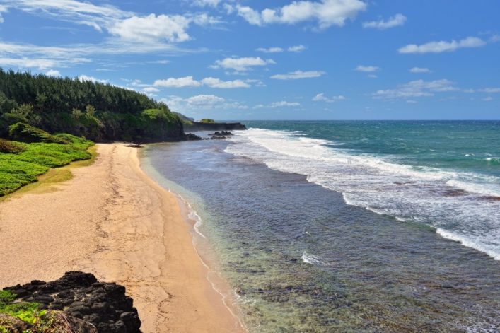 Weitläufiger Sandstrand links, umgeben von Klippen und üppigem Grün, daneben das blaue Meer mit sanften Wellen rechts. Am Horizont treffen Himmel und Meer aufeinander, unter leicht bewölktem Himmel.