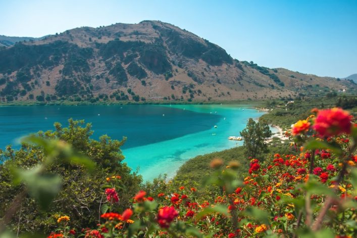 Türkisblauer Kournas-See auf Kreta, umringt von Bergen, mit blühenden Blumen im Vordergrund und einer Küstenlinie mit Bäumen.