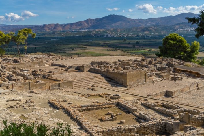 Antike Ruinen des minoischen Palastes von Phaistos auf Kreta, umgeben von Bergen und grüner Landschaft. Im Vordergrund Treppen und Mauern, im Hintergrund Hügel.