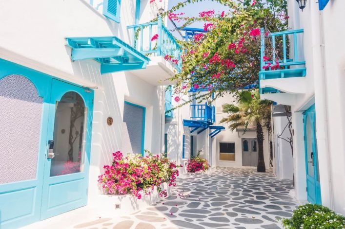 Schmale, gepflasterte Gasse in Mykonos mit weiß gestrichenen Häusern und blauen Türen und Balkonen. Pinke Blumen ranken über den Balkonen. Eine Palme steht an der rechten Seite der Gasse.