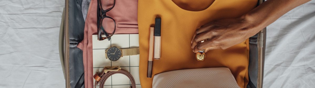 Ein halb geöffneter Koffer liegt auf einem weißen Bett. Darin Kleidung, eine Brille, eine Uhr, Gürtel, Schminke, eine weiße Tasche und eine Hand greift nach einem orangefarbenen Stoffstück.
