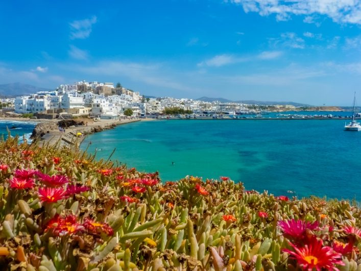 Bunte Blumen im Vordergrund, dahinter türkisfarbenes Meer mit einem Segelboot. Weiße Gebäude der Stadt Chora auf Naxos erheben sich links auf einer kleinen Anhöhe unter blauem Himmel.