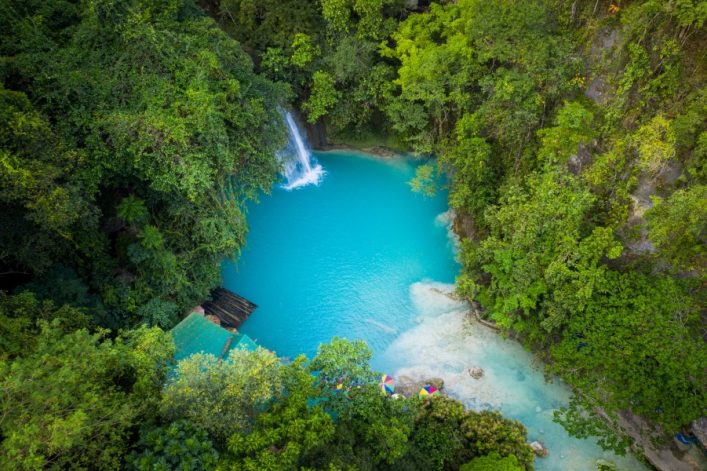 Türkisblauer Wasserfallpool inmitten dichter grüner Vegetation, Wasserfall links, von Bäumen und Sträuchern umgeben. Unten links ein Steg und bunte Sonnenschirme.