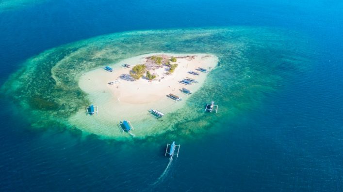 Kleine tropische Insel im türkisblauen Meer, umgeben von Sandbänken. Palmen und Vegetation im Zentrum, Boote umgeben die Insel. Klarer Himmel spiegelt sich auf dem Wasser.
