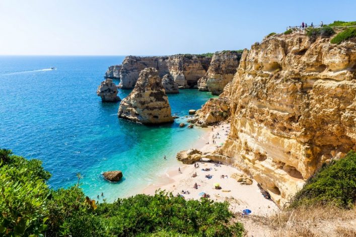 Sonniger Tag an der Algarve-Küste. Unten rechts ein goldener Sandstrand mit Sonnenanbetern und bunten Sonnenschirmen. Links das türkisfarbene Meer, dahinter majestätische Felsenklippen.