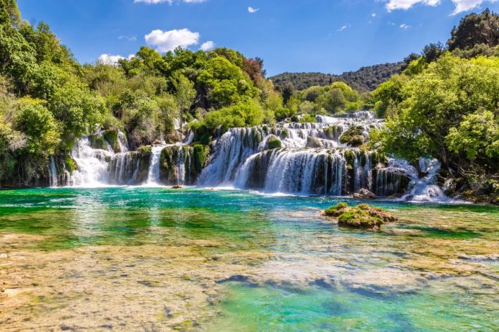 Wasserfall im Krka-Nationalpark, Kroatien: Mehrstufige Kaskaden, umgeben von üppigem Grün, fließen in smaragdgrünes Wasser. Im Hintergrund blauer Himmel und vereinzelte Wolken.