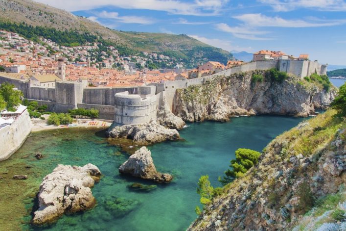 Stadtmauer von Dubrovnik, Kroatien, umgeben von klarem türkisfarbenem Meer. Häuser mit roten Dächern drängen sich dahinter. Im Vordergrund felsige Küste, im Hintergrund bewaldete Hügel.