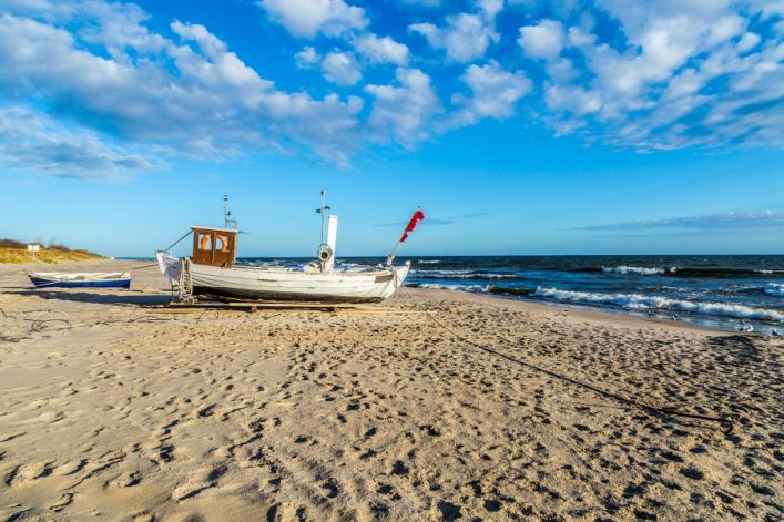 Ein weißes Fischerboot mit roten Flaggen liegt am hellen Sandstrand. Im Hintergrund das blaue Meer mit kleinen Wellen, darüber ein klarer Himmel mit einigen Wolken. Links im Bild Strandvegetation.