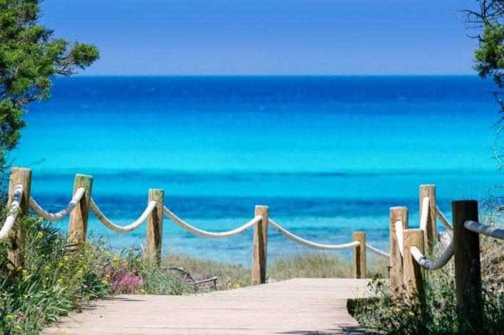 Holzsteg führt durch Dünen zum türkisblauen Meer. Links und rechts stehen Holzpfosten mit Seilen, umgeben von grüner Vegetation. Der Ozeanhorizont erstreckt sich bei klarem, blauem Himmel.