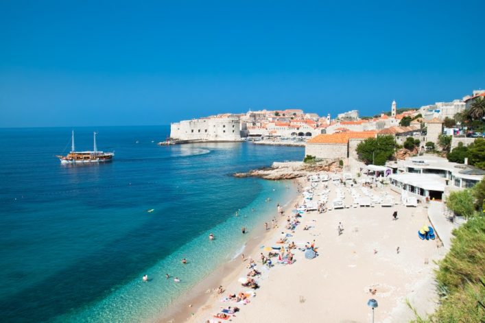 Strand mit hellem Sand, türkisblauem Meer links und zahlreichen Menschen. Im Hintergrund historische Stadtmauer von Dubrovnik, orangefarbene Dächer, rechts Bars und Sonnenschirme.