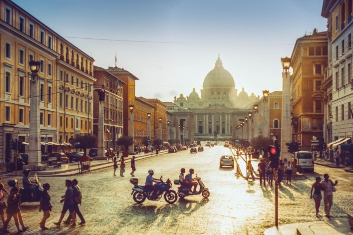 Petersdom im Hintergrund bei Sonnenuntergang, flankiert von historischen Gebäuden. Menschen gehen und fahren auf einer breiten Straße, einige auf Rollern und Motorrädern.