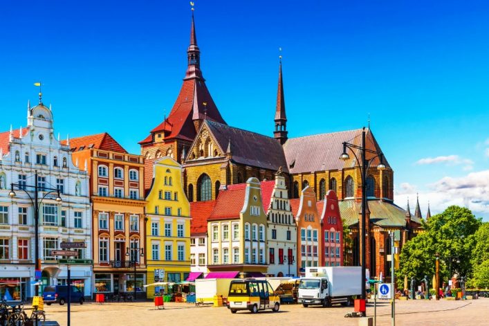 Bunte historische Häuser mit Giebeldächern in Rostock, darunter gelb, rot und orange, erstrecken sich von links nach rechts. Dahinter ragt eine große Kirche mit Türmen in den blauen Himmel.